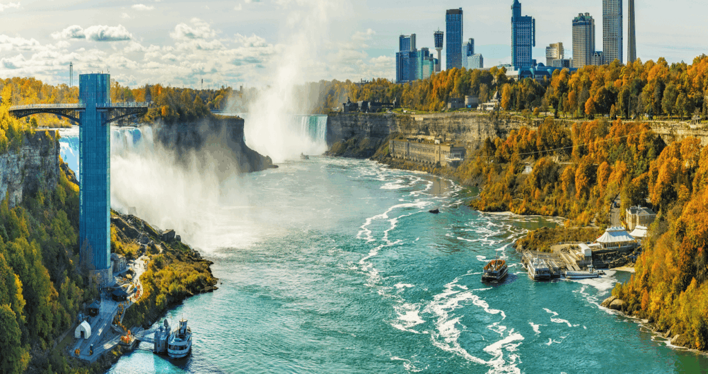 Panoramic view of Niagara Falls with water mist, vibrant autumn foliage, boats on the river providing cross-border services, and city buildings in the background.