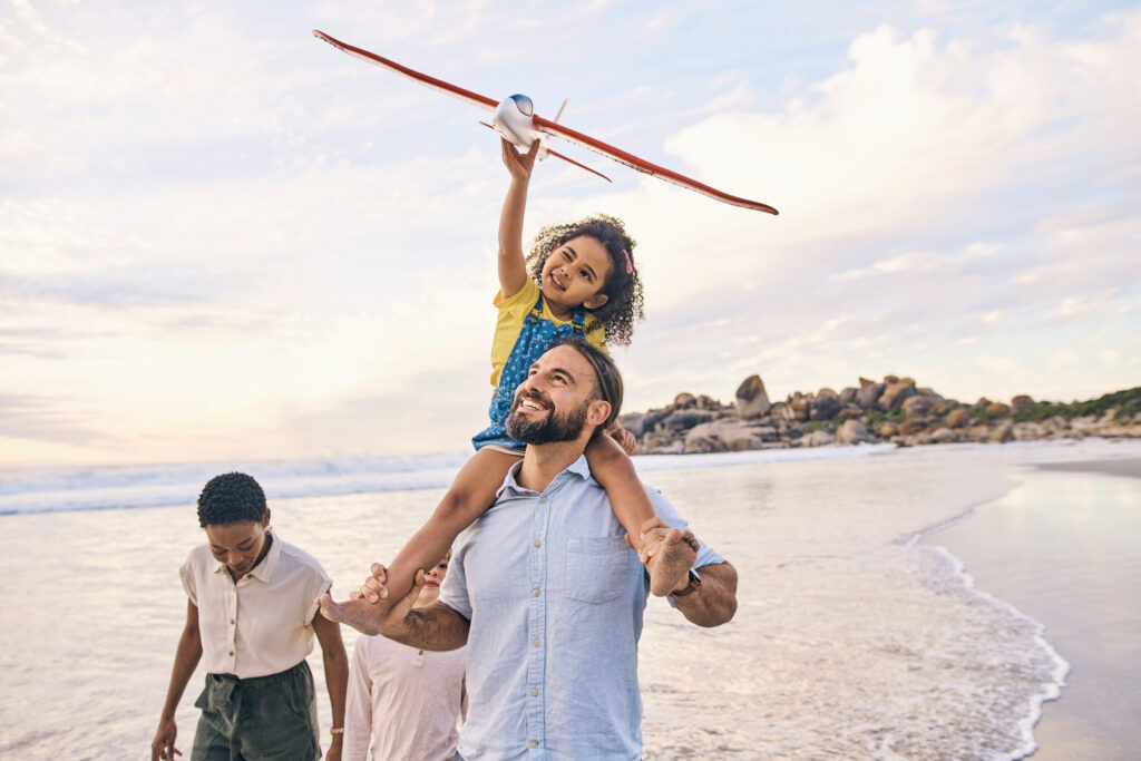 A child sits on an adult’s shoulders holding a toy airplane, walking with two others along a beach at sunset—capturing the joy and promise of generational wealth transfer.