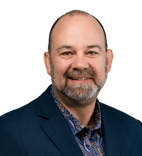 Smiling middle-aged man with a short beard, wearing a blue blazer and patterned shirt, facing the camera against a plain background.