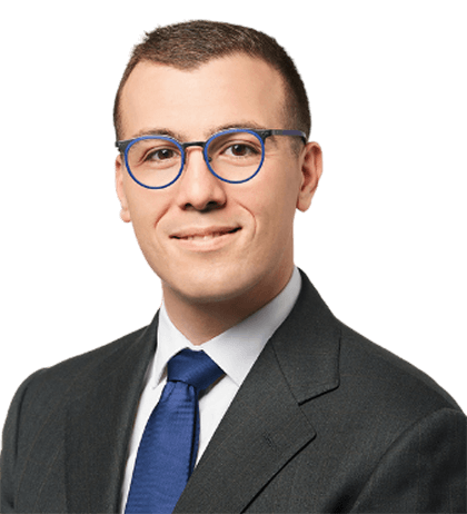 A man wearing blue-rimmed glasses, a dark suit, white shirt, and blue tie, posed against a plain background.