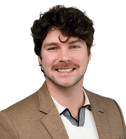 A man with curly brown hair and a mustache smiles, wearing a tan blazer over a white collared shirt, resembling Teddy Edward Leslie-MacDonald, against a plain background.