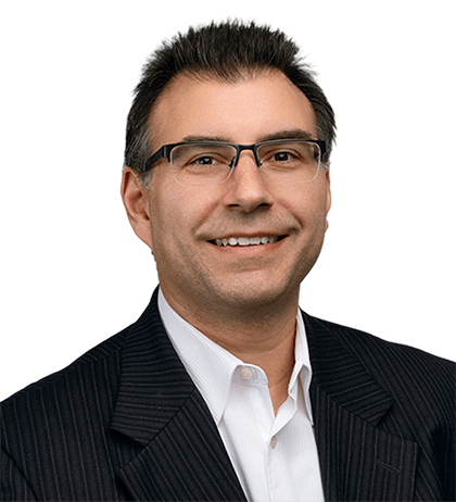 Eli Papakirykos, a man with short dark hair, glasses, and a pinstripe suit jacket over a white collared shirt, smiles at the camera against a plain background.