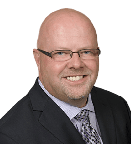 A bald man wearing glasses, a dark suit, and a patterned tie, smiling at the camera against a transparent background.