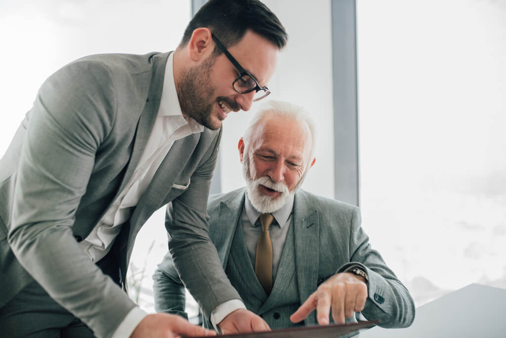 Two men in suits, one older and one younger, are looking at documents together and discussing succession planning in a bright office setting.
