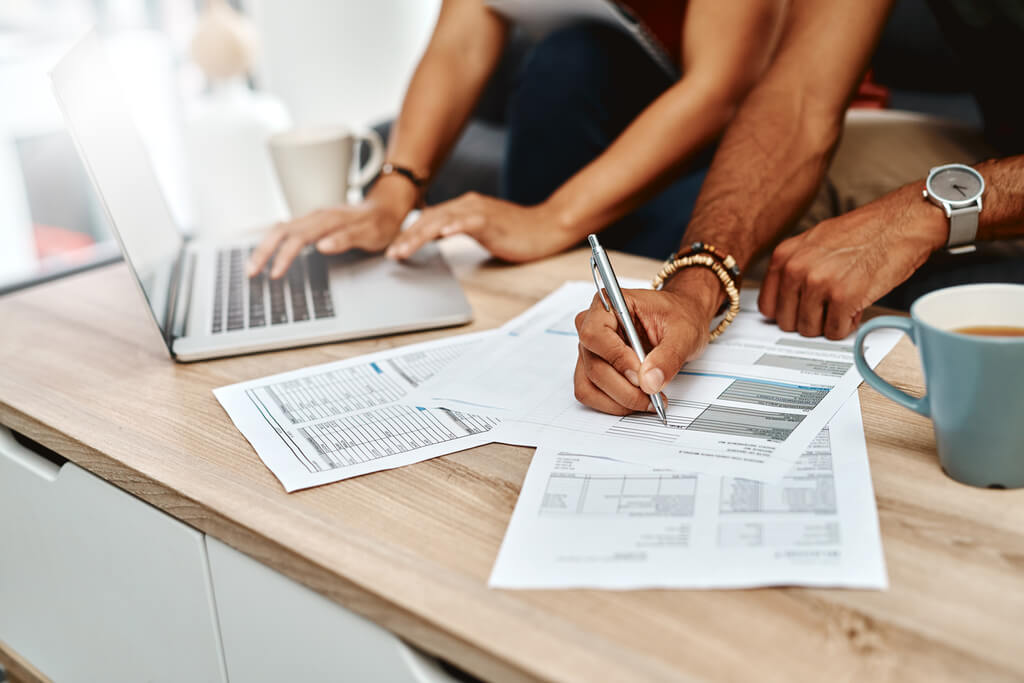 Two people work at a table with a laptop, coffee cup, and financial documents; one is writing while the other types on the laptop, collaborating on tax planning.