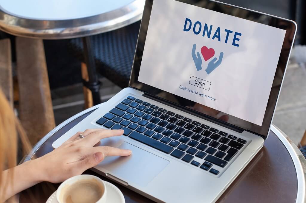 A person using a laptop with a screen displaying a donations webpage, featuring the word DONATE, two hands, a heart, and a send button—promoting charitable giving online.