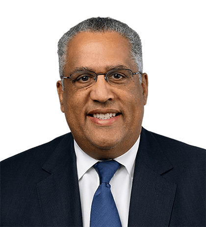 A man wearing glasses, a dark suit, white shirt, and blue tie, smiling at the camera against a plain background.