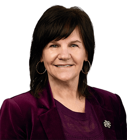 Woman with medium-length dark hair wearing a purple blazer, purple top, and hoop earrings, smiling at the camera against a transparent background.