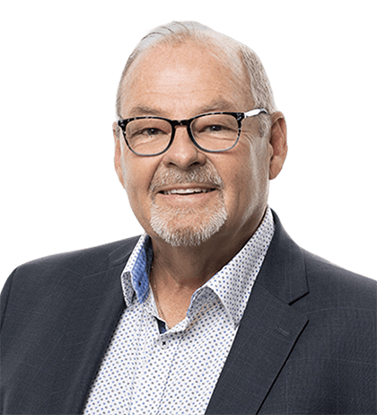 An older man with short gray hair, glasses, and a trimmed beard, wearing a dark suit jacket over a white patterned shirt, smiling at the camera.