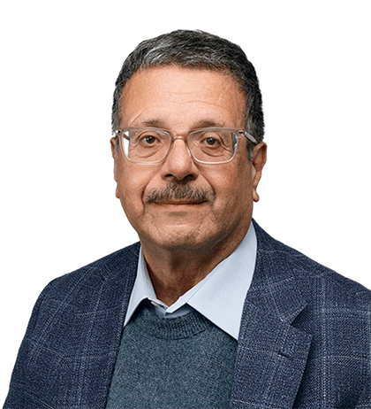 Man with short gray hair, glasses, and mustache, resembling Naguib Kerba, wearing a blue blazer, light blue shirt, and dark sweater, facing forward against a transparent background.