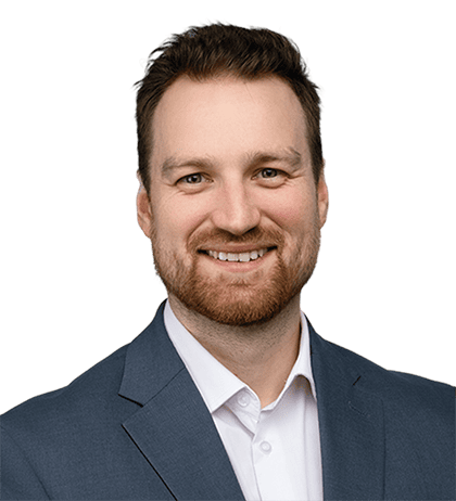 A man with short brown hair and a beard is smiling, wearing a blue suit jacket and a white collared shirt against a plain background.
