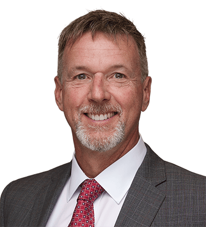 Richard Heinrich, a man with short gray hair and a goatee, wears a gray suit, white shirt, and red patterned tie, smiling at the camera against a plain background.
