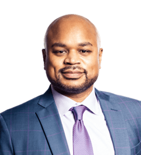 Richard Mitchell, wearing a blue checked suit, light-colored shirt, and purple tie, faces forward and smiles slightly.