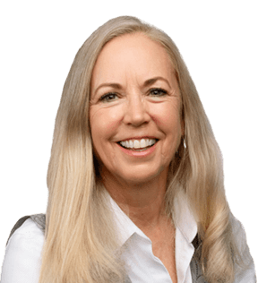 Susan Schulze, a woman with long blonde hair, wears a white blouse and gray vest, smiling at the camera against a plain background.
