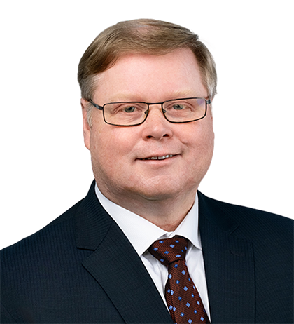 Will Beacham, a middle-aged man with light hair and glasses, is wearing a dark suit, white shirt, and patterned tie, posing against a blurred background.