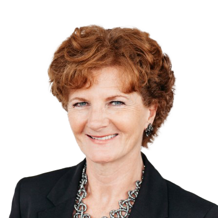 A woman with short curly reddish-brown hair, wearing a dark blazer, necklace, and earrings, smiling at the camera against a plain white background.