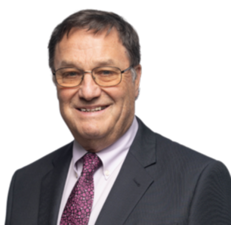 Ralph Alexander, a man in a suit, glasses, and a patterned tie, smiles at the camera against a plain, light background.