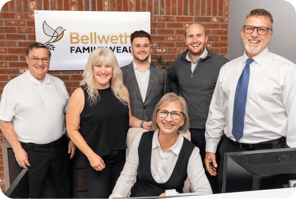 Six people, four men and two women, pose and smile in an office in front of a Bellwether Family Wealth sign against a brick wall—ready to help you take your next steps toward a confident retirement plan.
