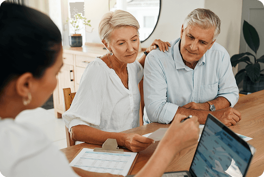 A couple sits at a table reviewing documents with a professional, who is pointing at a laptop screen as they discuss retirement planning. Papers and a calculator are visible on the table.